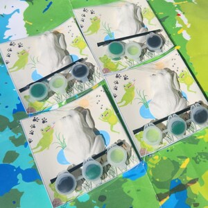 May include: Four white ceramic frog figurines with green paint pots and paintbrushes. Each frog is on a card with a green frog illustration and the words "Froggy Fun".