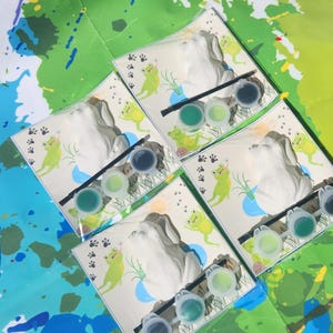 May include: Four sets of frog-themed paint-your-own ceramic figurines. Each set includes a white ceramic frog figurine, a small paintbrush, and four small pots of green paint.