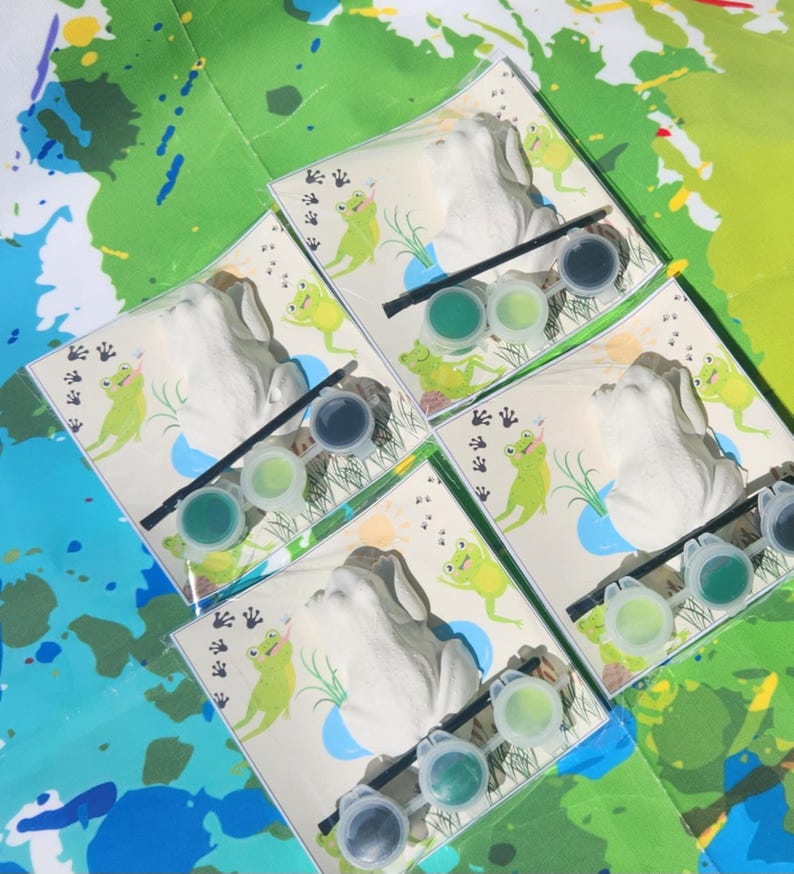 May include: Four sets of frog-themed paint-your-own ceramic figurines. Each set includes a white ceramic frog figurine, a small paintbrush, and four small pots of green paint.