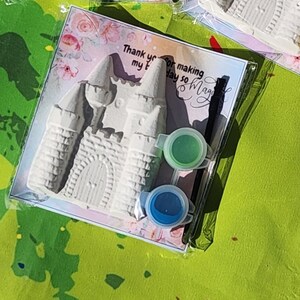 May include: A paint-your-own castle kit in a clear plastic package. The kit includes a gray castle, two paint pots with green and blue paint, and a card with text that reads "Thank you for making my day so magic".