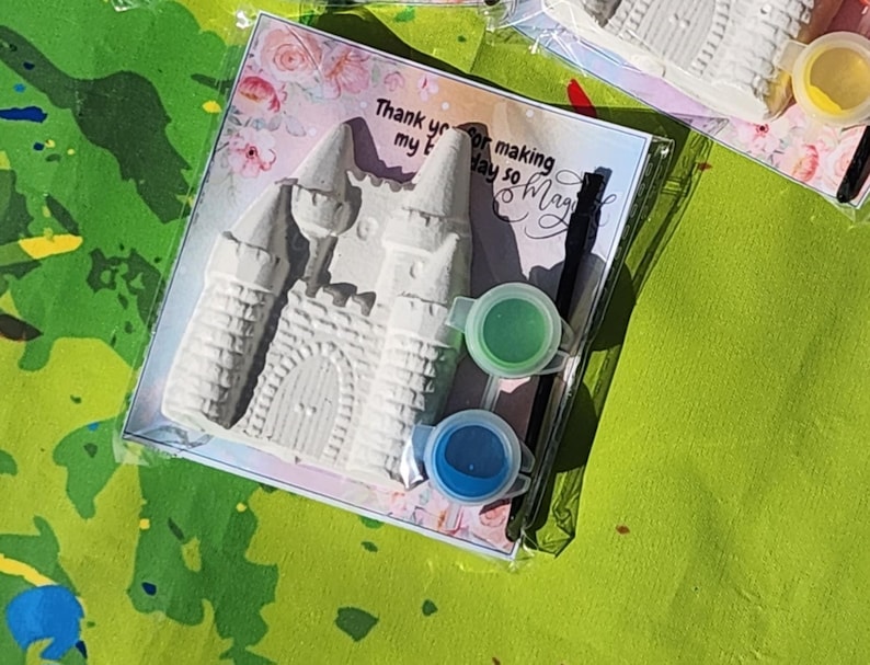 May include: A paint-your-own castle kit in a clear plastic package. The kit includes a gray castle, two paint pots with green and blue paint, and a card with text that reads "Thank you for making my day so magic".
