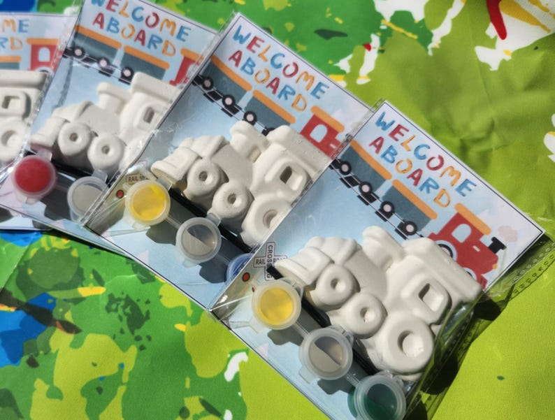 May include: A set of three paint-your-own ceramic train engine ornaments with the words "Welcome Aboard" printed on each. The set includes paint pots in red, yellow, green, and white.