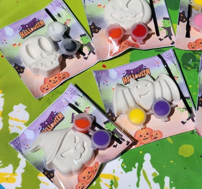 May include: A set of four Halloween-themed paint-your-own kits. Each kit includes a white ceramic figurine, a small paintbrush, and three small pots of paint. The figurines are a skull, a pumpkin, a bat, and a ghost. The kits are packaged in clear plastic bags with a Happy Halloween label.