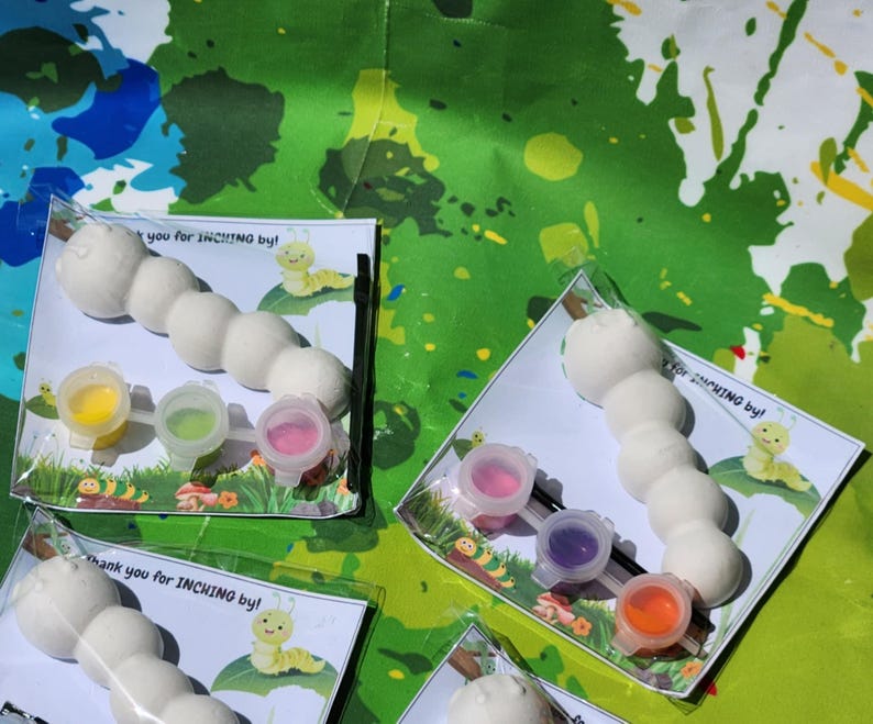 Może przedstawiać: Zestaw czterech zestaw&oacute;w do samodzielnego malowania gąsienic. Każdy zestaw zawiera białą ceramiczną gąsienicę, cztery małe pojemniki z farbą i pędzel. Pojemniki z farbą są ż&oacute;łte, zielone, r&oacute;żowe i fioletowe. Zestawy są zapakowane w przezroczyste plastikowe torebki z podziękowaniem za pełzanie!