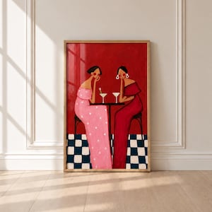 May include: A framed art print featuring a painting of two figures seated at a small table against a solid red background. One figure wears a pink polka-dot dress, the other a solid red dress. They sit on a black and white chequered floor.