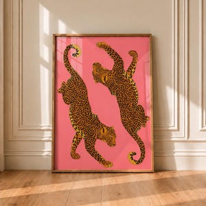 Könnte beinhalten: Ein gerahmter, rechteckiger Wandkunstdruck mit zwei Leoparden mit orangefarbenem, schwarz geflecktem Fell vor einem einfarbigen rosa Hintergrund. Die Leoparden sind in dynamischen Posen dargestellt, einer springend und einer gehend, in einem dünnen Holzrahmen.