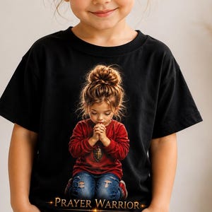 Op de afbeelding: Een zwart t-shirt met korte mouwen en een grafische print van een kind met een rommelige knot, gekleed in een rood shirt met lange mouwen en een blauwe spijkerbroek, knielend met gevouwen handen. De tekst PRAYER WARRIOR staat in goud eronder.