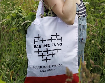 Könnte beinhalten: Eine weiße Tragetasche mit einem roten unteren Rand. Auf der Vorderseite befindet sich eine schwarze Grafik in Herzform, die aus kleinen Kreuzen besteht, mit dem Text "BAG THE FLAG" darüber und "TOLERANCE PEACE AND UNITY" darunter.