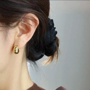 Puede incluir: Un pendiente pequeño de tono dorado pulido con una forma suave, redondeada y similar a una gota. El pendiente presenta una superficie metálica reflectante y está sujeto al lóbulo de la oreja. El diseño es simple, compacto y brillante.