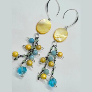 Può includere: Un paio di orecchini pendenti con cabochon ovali in madreperla gialla fissati a ganci argentati. Sotto ogni cabochon, diversi fili di metallo scuro sostengono piccole perline e cristalli di colore giallo, azzurro e verde pallido.