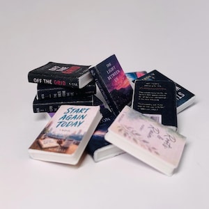 May include: A collection of miniature books with various titles, including "Off the Grid" and "Start Again Today." The books have colorful covers and are stacked and scattered against a white background. The books are small, likely for a dollhouse or display.