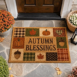May include: A rectangular coir doormat featuring a patchwork design with the text AUTUMN BLESSINGS in the center. The border includes illustrations of orange pumpkins, brown acorns, red maple leaves, and various plaid and polka dot patterns.