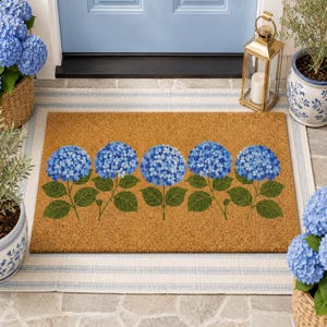 May include: A rectangular natural tan coir doormat featuring a horizontal row of five vibrant blue hydrangea flower clusters with green leaves printed across the center. The mat has a coarse, fibrous texture typical of coconut husk material.