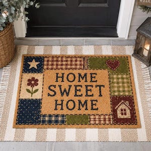 May include: A rectangular coir doormat featuring a central tan panel with the black text HOME SWEET HOME. The border consists of patchwork squares in navy blue, olive green, and brown plaid, decorated with a star, flower, heart, and house.