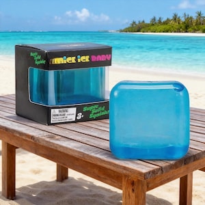 May include: A blue, squishy toy cube with rounded edges sits on a wooden table. The toy is next to its black box, which reads "Nice Ice Baby" and "Super Solid Squish!" The background shows a beach and ocean.