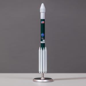 May include: A model rocket with a white nose cone and a dark green body. The body features the NASA logo, the American flag, and other details. The rocket is mounted on a silver base.