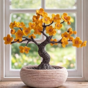 Könnte beinhalten: Ein handgefertigter Bonsai-Baum mit leuchtend gelben gehäkelten Blüten. Der Baum hat einen gedrehten, grauen Stamm und Äste, die in einem gewebten, hellbeigen Topf stehen. Der Hintergrund zeigt ein Fenster mit verschwommenem Grün.