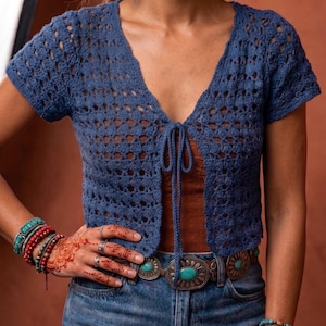 Könnte beinhalten: Ein kurzärmliges, offenes, blaues Häkeltop mit Kordelverschluss. Das Top wird mit einem braunen Tanktop, einer Jeans und einem Gürtel mit Türkis-Akzenten kombiniert. Das Model trägt außerdem Armbänder und Ringe.