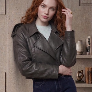 May include: A dark gray leather jacket with a classic collar and a double-breasted design. The jacket is paired with dark blue denim jeans. The model is wearing a light blue sweater underneath the jacket.