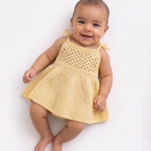 Op de afbeelding: Een lichtgele gehaakte babyjurk met een mouwloos ontwerp en strikbandjes. De jurk heeft een getextureerd lijfje en een uitlopende rok. De baby ligt op een witte achtergrond.
