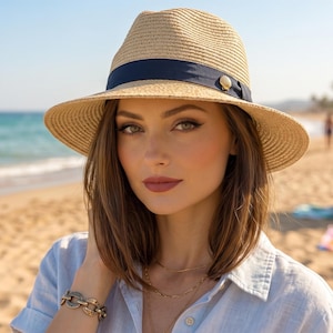 May include: A straw fedora hat with a navy blue band and a decorative button. The hat is worn by a person on a beach. The background includes the ocean, sand, and palm trees, suggesting a sunny day.