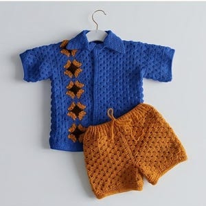 May include: A blue crochet shirt and shorts set. The short-sleeved shirt has a collar and is decorated with brown diamond shapes. The shorts are a matching orange colour with a drawstring waist. The set is displayed on a white hanger.