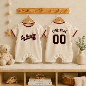 Könnte beinhalten: Zwei cremefarbene Baby-Strampler mit kurzen Ärmeln und orange-schwarzen Abschlüssen. Die Vorderseite eines Stramplers zeigt den Schriftzug San Francisco mit einer Baseball-Grafik. Die Rückseite des anderen zeigt den Text YOUR NAME und die Zahl 00.