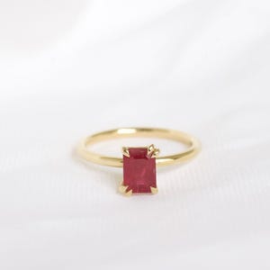 Peut inclure: Une bague en or ornée d'une pierre rouge rectangulaire taille émeraude fixée par quatre griffes. L'anneau est fin, lisse et poli, présentant un design épuré qui met en valeur la pierre rouge vive comme point focal central de la pièce.