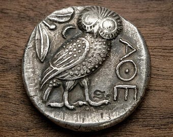 May include: A silver coin featuring a detailed owl with olive branch and the Greek letters ΑΘΕ. The coin has a worn, antique appearance, with visible signs of age and use. The owl is the central focus, with intricate detailing on its feathers and face.