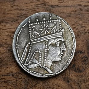 May include: A silver coin featuring a profile of a person wearing a crown and a detailed border. The coin's design includes intricate patterns and symbols, suggesting historical significance. The coin is displayed on a wooden surface.