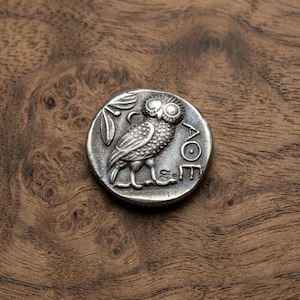 May include: A silver coin featuring a detailed owl design, with the Greek letters 'AOE' visible. The coin is round and has a slightly worn appearance, suggesting age. The owl is depicted with intricate details, including feathers and large eyes. The coin is set against a wooden background.