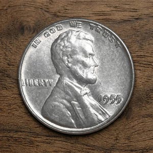 以下が含まれることがあります： 1955年のリンカーンペニー銀貨。エイブラハム・リンカーンの横顔、"LIBERTY"の文字、"IN GOD WE TRUST"のフレーズが特徴です。木製の背景に置かれています。