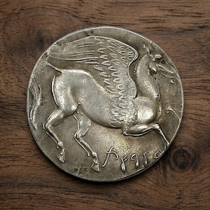May include: A silver coin featuring a detailed relief of a winged horse, likely Pegasus, in mid-flight. The coin has an aged appearance, with visible wear and tear. The background is a dark wooden surface, providing contrast to the coin's metallic sheen.