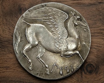 May include: A silver coin featuring a detailed relief of a winged horse, likely Pegasus, in mid-flight. The coin has an aged appearance, with visible wear and tear. The background is a dark wooden surface, providing contrast to the coin's metallic sheen.