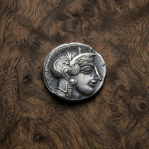 May include: A silver coin featuring a detailed profile of a woman with ornate headwear and jewelry. The coin is set against a dark, textured wooden surface, highlighting its intricate design and historical aesthetic.