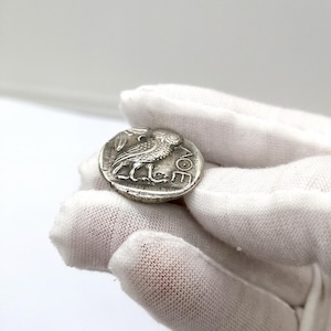 May include: A silver coin featuring an owl with a sprig of leaves. The coin has ancient Greek lettering and is held in a gloved hand. The coin is round and has a detailed relief design.