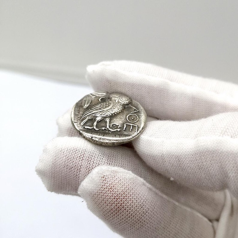May include: A silver coin featuring an owl with a sprig of leaves. The coin has ancient Greek lettering and is held in a gloved hand. The coin is round and has a detailed relief design.