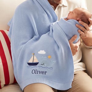 Peut inclure: Une couverture pour bébé en tricot bleu clair ornée d'un voilier, d'une baleine souriante, d'un soleil et d'un nuage brodés. Le prénom Oliver est cousu en fil bleu foncé sous le motif nautique, avec une bordure côtelée sur le bas.