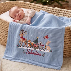 Puede incluir: Una manta de bebé tejida en color azul claro con un bordado que muestra un ciervo, un zorro, un mapache y un conejo junto a un árbol de Navidad y regalos. El nombre Sebastian está bordado en color rojo debajo de los animales con acebo.