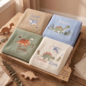 May include: Four folded knit blankets in tan, light blue, sage green, and cream, each featuring embroidered dinosaur motifs and personalised names. The cream blanket displays the names Emma and Liam, while the others are personalised with Jason.