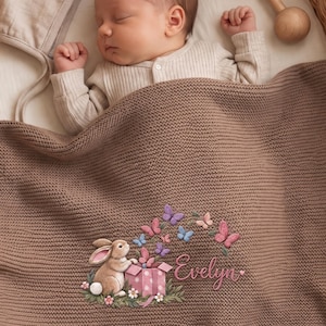 May include: A brown knitted baby blanket featuring an embroidered design of a light brown rabbit sitting next to a pink polka-dot gift box with colourful butterflies flying above it and the name Evelyn written in pink cursive script lettering.