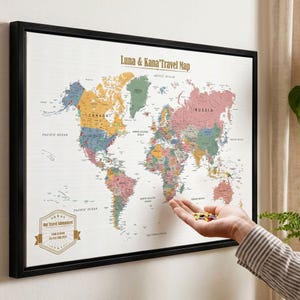 May include: A framed world map with a white background and multicolored continents. The text reads Luna & Kana's Travel Map and Our Travel Adventures Luna & Kana 08. Feb 10th 2025. Small colorful pins are held in a hand near the map surface.
