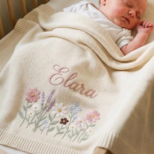 Könnte beinhalten: Eine cremefarbene Strickdecke für Babys mit dem eingestickten Namen Elara in einer hellrosa Schreibschrift. Unter dem Namen befindet sich ein detailliertes Blumenmuster mit gestickten Blüten in Rosa, Weiß, Blau und Braun mit grünen Stielen.