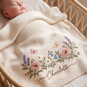 May include: A cream-colored knit baby blanket featuring an embroidered floral arrangement with pink, white, blue, and purple flowers, green leaves, and two butterflies. The name Charlotte is embroidered in brown cursive script on the blanket.
