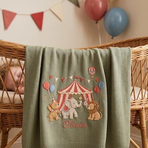 Può includere: Una coperta per neonati in maglia verde salvia con una scena circense ricamata che include un tendone a righe rosse e bianche, un orso, un elefante, un leone e dei palloncini. Il nome Olivia è ricamato in corsivo rosso sotto gli animali.