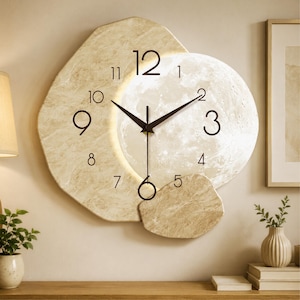 Peut inclure: Une horloge murale beige avec un design en forme de lune. Le cadran de l'horloge comporte des chiffres et des aiguilles noirs, avec une lune lumineuse au centre. L'horloge mesure environ 46 cm de diamètre.