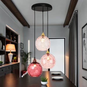 Può includere: Tre lampade a sospensione con globi di vetro rosa e dettagli dorati pendono da un apparecchio a soffitto nero. I globi hanno una superficie strutturata e variano nelle tonalità di rosa, creando un'atmosfera calda. Le lampade sono sospese sopra un tavolo di legno scuro.