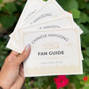 Peut inclure: Un paquet de cartes rectangulaires blanches intitulées Chinese Mahjong Fan Guide Hong Kong Style. Les cartes présentent du texte noir, des bordures dorées et des illustrations de trois tuiles de mahjong au centre avec des vagues en bas.