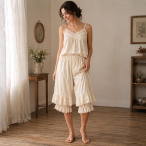 May include: Cream-colored pajama set featuring a camisole top with lace trim and matching cropped pants with ruffled hems. The soft, light-colored fabric creates a comfortable and elegant sleepwear ensemble. The set is designed for a relaxed fit.