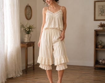May include: Cream-coloured pajama set featuring a camisole top with lace trim and matching cropped trousers with ruffled hems. The soft, light-coloured fabric creates a comfortable and elegant sleepwear ensemble. The set is designed for a relaxed fit.